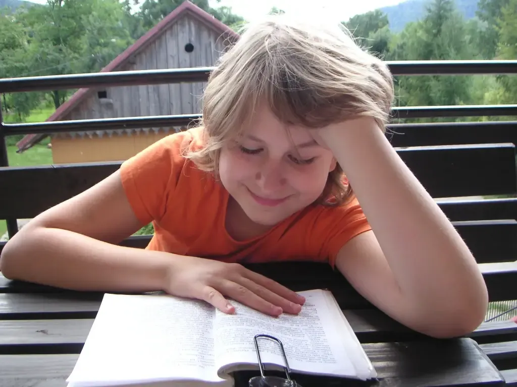 Dziewczynka siedzi przy stole na tarasie i z uśmiechem odrabia lekcje.